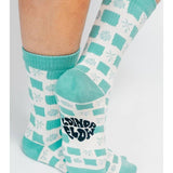 A person wearing the Tropical Checkers Knit Crew Sock by Tailored Union—teal and white checks with palm tree designs, made from premium combed cotton, and "SOUND & COLOR" printed on the sole.