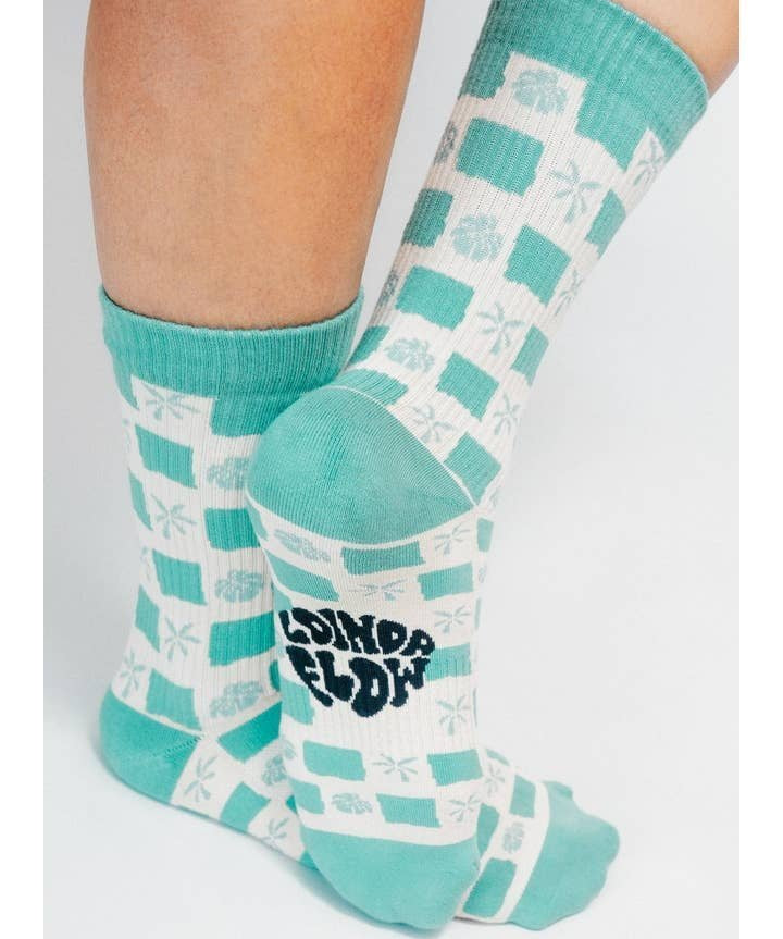 A person wearing the Tropical Checkers Knit Crew Sock by Tailored Union—teal and white checks with palm tree designs, made from premium combed cotton, and "SOUND & COLOR" printed on the sole.