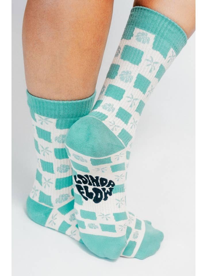 A person wearing the Tropical Checkers Knit Crew Sock by Tailored Union—teal and white checks with palm tree designs, made from premium combed cotton, and "SOUND & COLOR" printed on the sole.