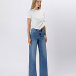 A woman stands against a plain background wearing a white t-shirt, silver high-heeled sandals, and Vervet's Tummy Control Super High Rise Baggy Wide Jeans in blue for a relaxed vibe.