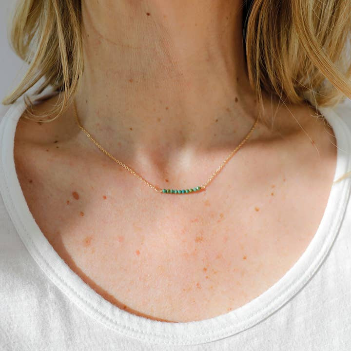 A person wears the Turquoise Bead Necklace by Ornamental Things, featuring a thin gold filled chain with small turquoise beads, visible on their neck and upper chest above a white shirt.