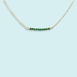 The Turquoise Bead Necklace by Ornamental Things features a gold-filled chain with a horizontal row of small turquoise beads, shown against a light blue background.