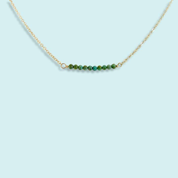 The Turquoise Bead Necklace by Ornamental Things features a gold-filled chain with a horizontal row of small turquoise beads, shown against a light blue background.