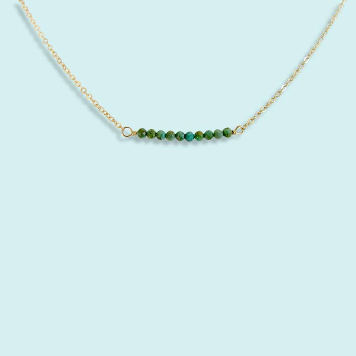 The Turquoise Bead Necklace by Ornamental Things features a gold filled chain and a row of small turquoise beads, displayed on a light blue background. This adjustable piece adds elegant charm to any outfit.