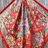 The Rodeo Drive "Turquoise That's So 70's Paisley Wild Rag" is a square silk scarf with vibrant floral and paisley designs on a turquoise background, displayed flat on a wooden surface.