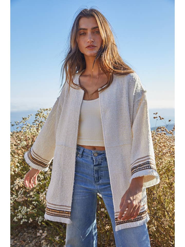 A woman stands outdoors in a cream Tweed Long Sleeve Jacket by By Together, featuring striped cuffs, long sleeves, and blue jeans, with plants and a blue sky in the background.