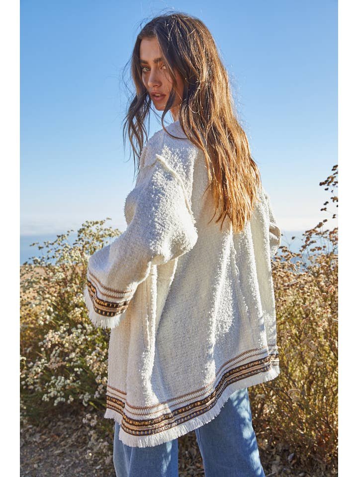 A woman with long brown hair stands outdoors against wildflowers and a blue sky, wearing the By Together Tweed Long Sleeve Jacket paired with blue jeans.
