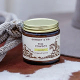 A jar of Cowboy & Co. Unscented Whipped Tallow Moisturizer sits on a surface next to a rope, with cowboys and horses on the label, highlighting grass-fed tallow as a key ingredient.