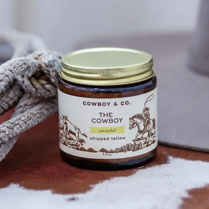 A jar of Cowboy & Co. Unscented Whipped Tallow Moisturizer sits on a surface next to a rope, with cowboys and horses on the label, highlighting grass-fed tallow as a key ingredient.