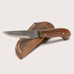 The Upland Game Knife by Woody Brand Knives, handmade in the USA with a wooden handle, sits on a brown leather sheath—ideal as an EDC personal knife.