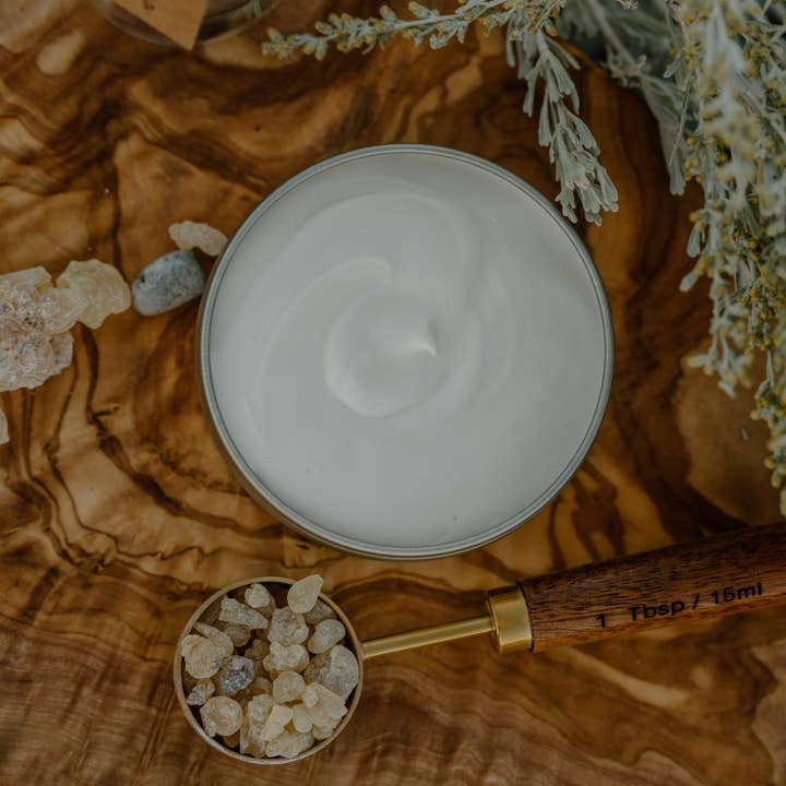 A tin of Cowboy & Co. Vanilla/Amber Nourishing Hydrating Tattoo Butter sits on a wooden surface beside resin pieces and greenery, ideal for tattoo aftercare.