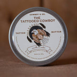 A tin of Cowboy & Co. Nourishing Hydrating Tattoo Butter with a label showing an illustrated eagle and text about its vanilla & amber scent, ingredients, and tattoo aftercare benefits.