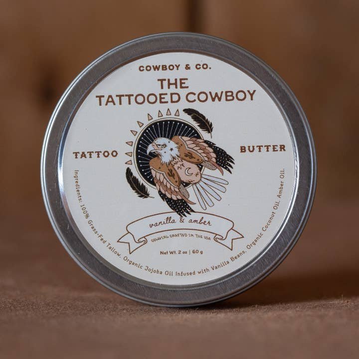 A tin of Cowboy & Co. Nourishing Hydrating Tattoo Butter with a label showing an illustrated eagle and text about its vanilla & amber scent, ingredients, and tattoo aftercare benefits.
