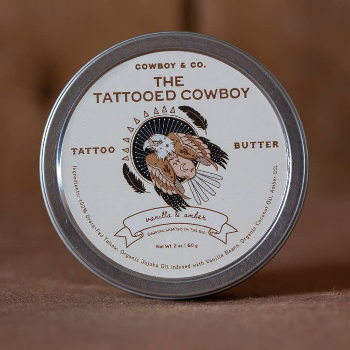 A round tin of Cowboy & Co. Vanilla/Amber Nourishing Hydrating Tattoo Butter, featuring an eagle illustration and ingredients listed around the edge—ideal for tattoo aftercare with a rich vanilla and amber scent.