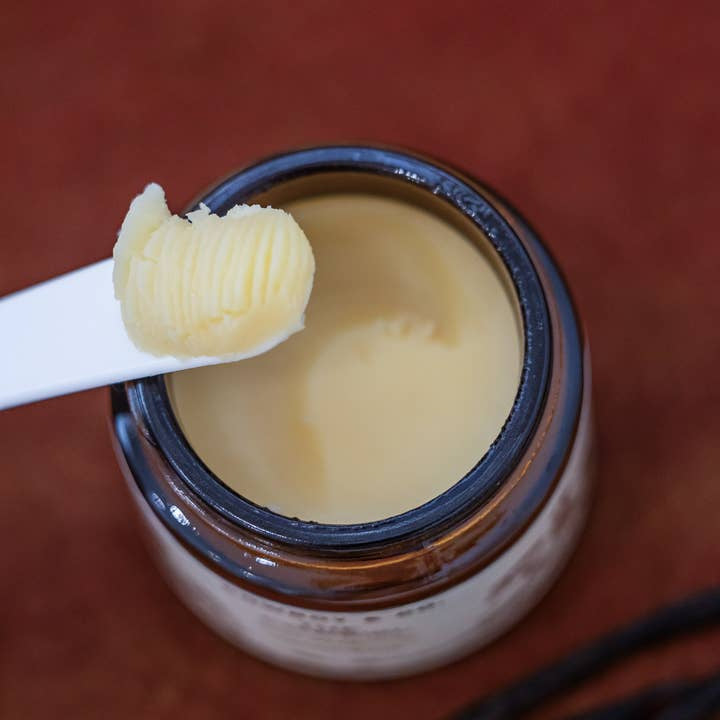 A close-up of an open jar of Vanilla Infused Tallow Balm by Cowboy & Co., with a spatula lifting a small amount above the jar, set against a reddish-brown background.