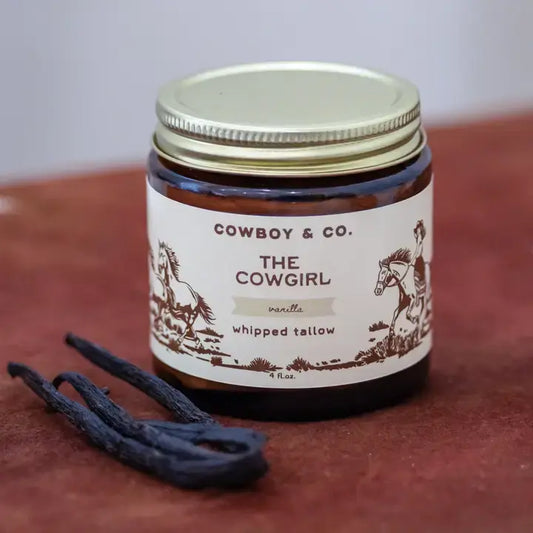 A glass jar labeled "Vanilla Whipped Tallow Moisturizer | Cowboy & Co." from Cowboy & Co. rests on a brown surface beside two vanilla bean pods.