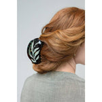 A woman with auburn hair in a low updo, secured with the Vervain Flower Herb Claw Clip by Winona Irene—a protective black clip featuring a white and green floral design.