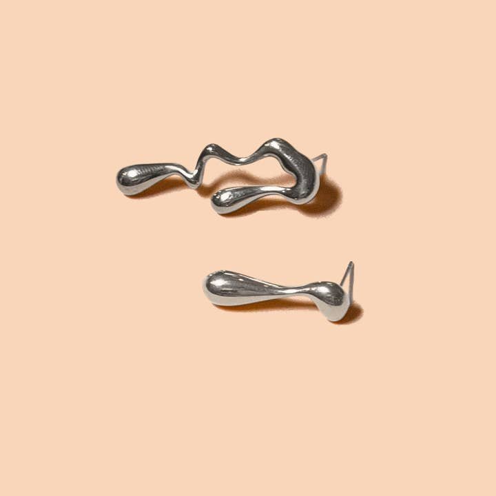 The Vice Versa Studs by Peter and June are two abstract, shiny silver earrings with fluid, irregular shapes crafted from waterproof stainless steel, displayed on a plain beige background.