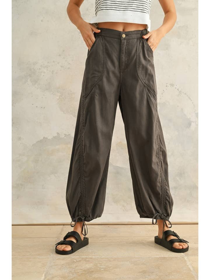 Wearing Miou Muse's Washed Drawstring On Bottom Pants—high-waisted, loose, charcoal tencel-cotton with cinched cuffs—paired with black sandals and a cropped striped top, standing against a neutral background.