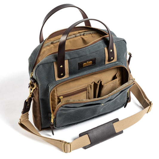 The Duck Head Waxed Canvas Laptop Bag in Charcoal features leather accents, multiple zippered compartments, brown handles, an adjustable beige shoulder strap, visible interior pockets, and a handy trolley sleeve for travel.