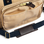 Close-up of the Duck Head Waxed Canvas Laptop Bag in beige, featuring a zippered pocket, brass hardware, and a navy blue adjustable shoulder strap with a brown leather pad. Made in the USA.