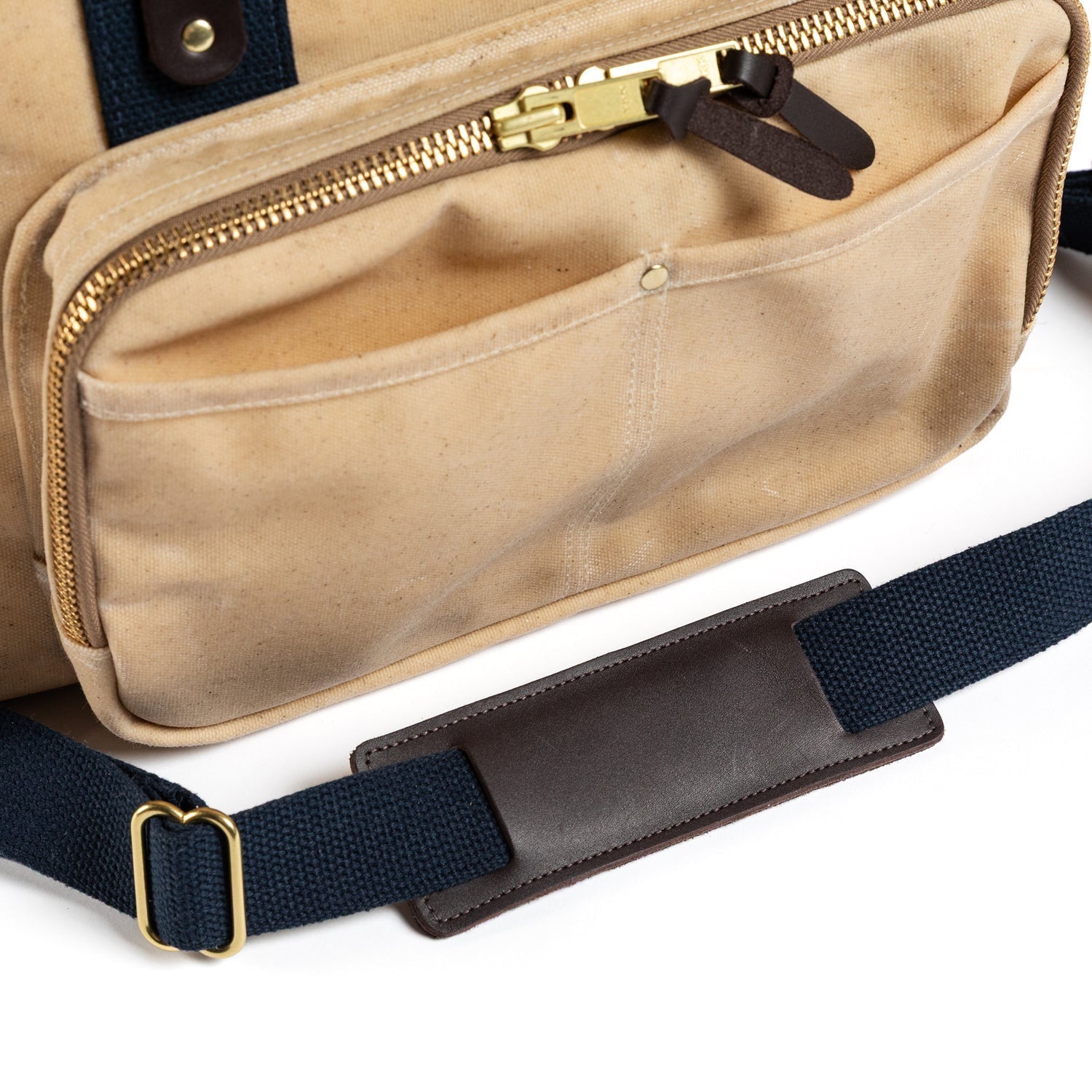 Close-up of the Duck Head Waxed Canvas Laptop Bag in beige, featuring a zippered pocket, brass hardware, and a navy blue adjustable shoulder strap with a brown leather pad. Made in the USA.