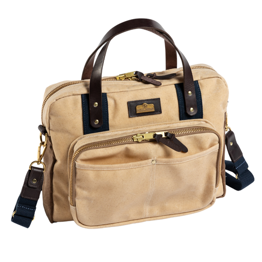 The Duck Head Waxed Canvas Laptop Bag features beige canvas, brown leather handles, gold hardware, a front zip pocket, detachable navy shoulder strap, and trolley sleeve—made in the USA and shown on a white background.