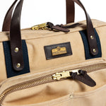 Close-up of the Duck Head Waxed Canvas Laptop Bag featuring tan waxed canvas, dark brown leather handles, blue fabric accents, gold zippers, and a visible "Duck Head" brand label on the front.