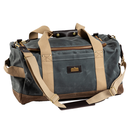 The Duck Head Waxed Canvas Overnighter Bag in charcoal features tan straps, gold-tone hardware, a front pocket, and a detachable shoulder strap. Made in the USA, it's your durable travel companion.