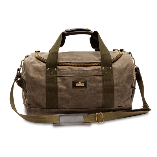The Duck Head Waxed Canvas Overnighter Duffel in Field Brown features olive green straps, a front zipper pocket, brass hardware, and a detachable shoulder strap, shown against a white background.