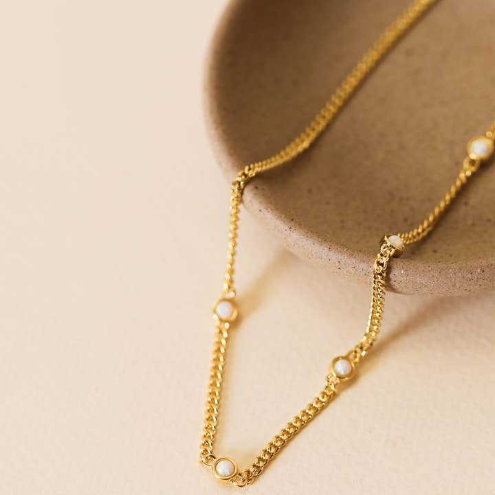 The JaxKelly White Opal Necklace—an 18k gold plated, hypoallergenic chain with small white opal stones—is displayed on the edge of a beige ceramic dish against a light background.