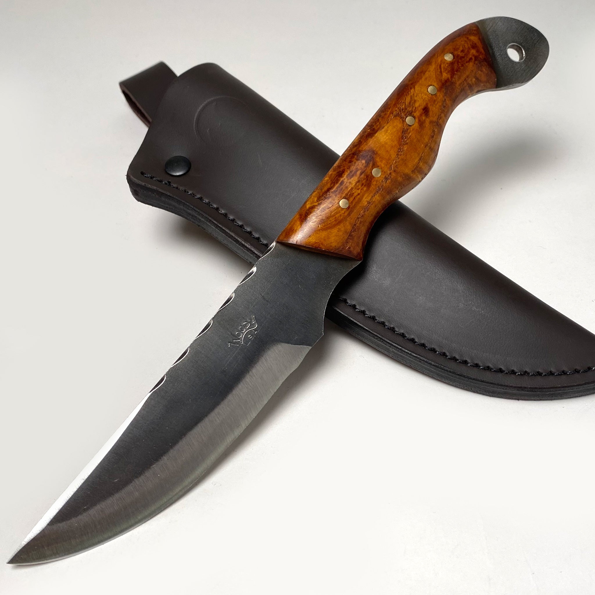 The White Skunk Camp Knife by Woody Brand Knives, featuring a polished wooden handle and 1095 high carbon steel blade, rests atop a dark leather sheath against a plain background.