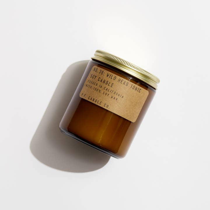 Amber jar candle with gold lid and brown paper label reading "Wild Herb Tonic | P.F. Candle Co." by P.F Candle, made with soy wax and paraben-free fragrance, photographed on a white background.