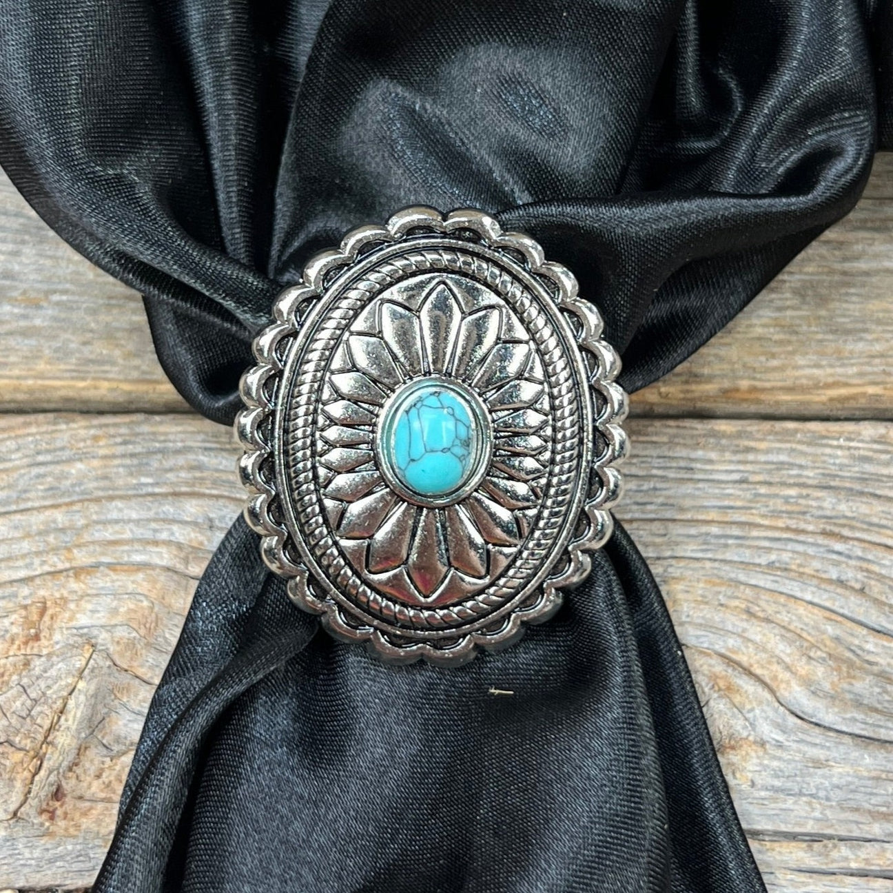 The Rodeo Drive Wild Rag Slide features an oval silver western concho with engraved patterns and a turquoise-colored stone center, shown on black fabric over a wooden surface.