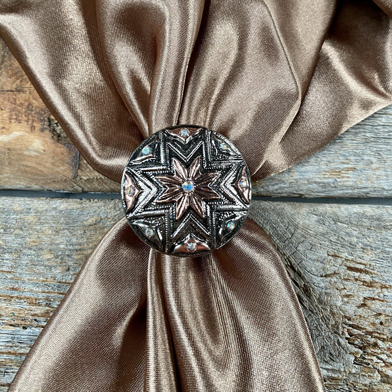 The Wild Rag Slide by Rodeo Drive features a round, ornate ring with star patterns and turquoise stone accents, shown holding a bronze satin napkin on a rustic wooden surface.