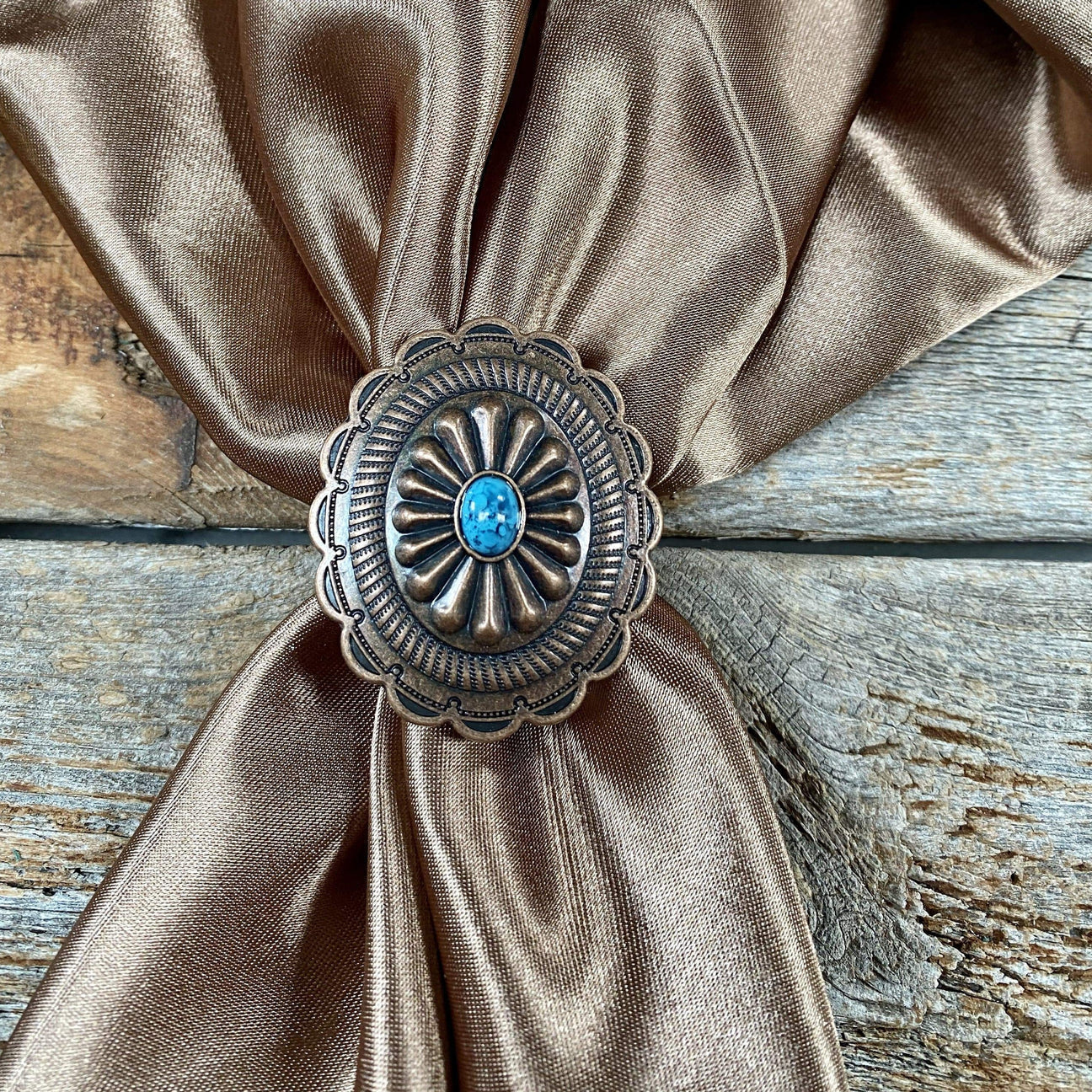 The Wild Rag Slide | Rodeo Drive by Rodeo Drive, featuring a bronze western concho with a turquoise-colored center, secures a brown satin wild rag displayed on weathered wood.