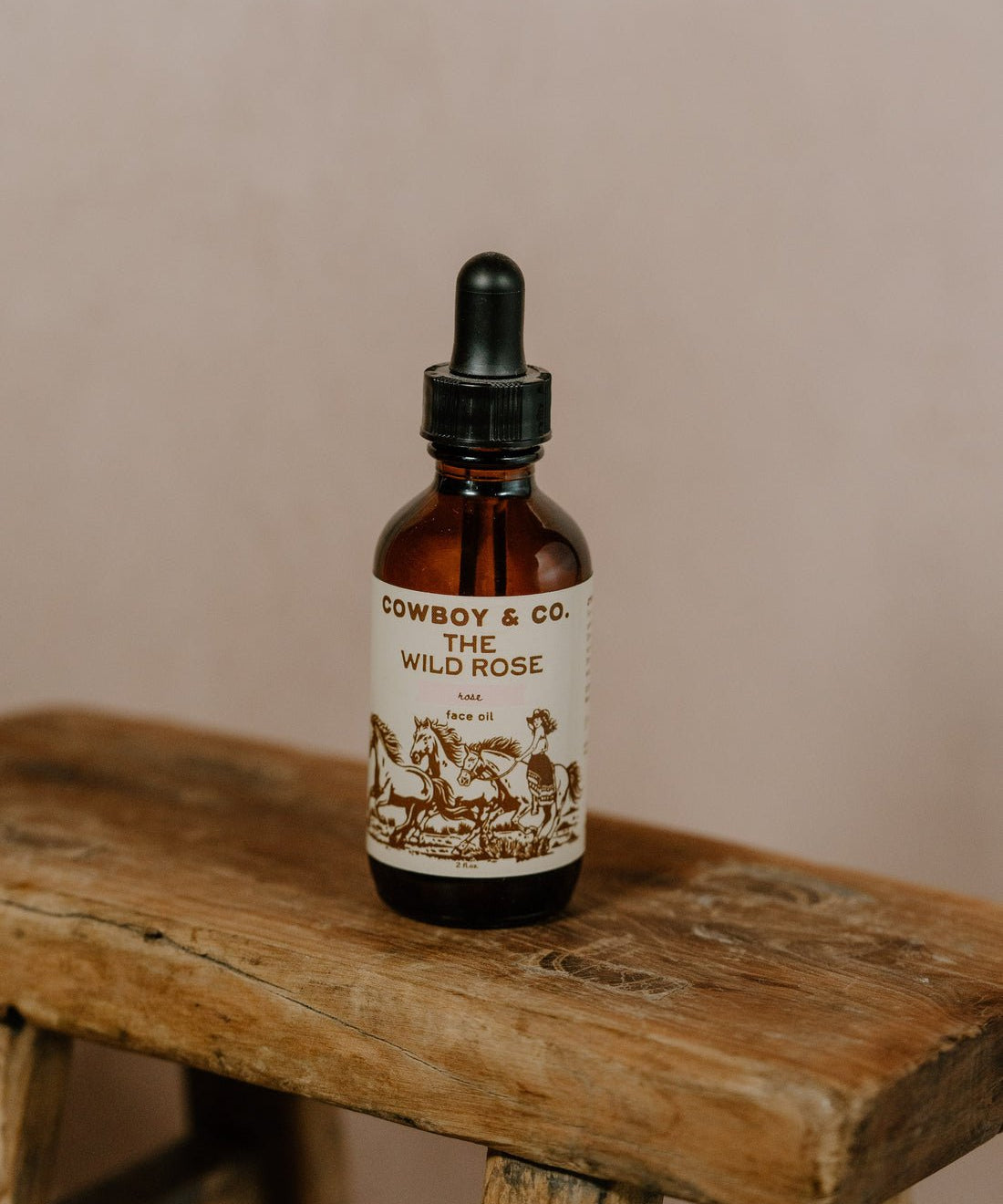 A brown glass dropper bottle labeled "WILD ROSE | Rose Infused Face Oil | Cowboy & Co." rests on a rustic stool, highlighting this hydrating face oil with organic rose petals by Cowboy & Co. against a neutral background.