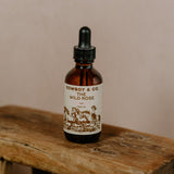 A brown glass dropper bottle labeled "WILD ROSE | Rose Infused Face Oil | Cowboy & Co." rests on a rustic stool, highlighting this hydrating face oil with organic rose petals by Cowboy & Co. against a neutral background.