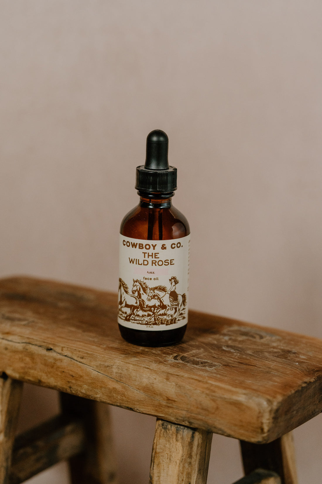 A brown glass dropper bottle labeled "WILD ROSE | Rose Infused Face Oil | Cowboy & Co." rests on a rustic stool, highlighting this hydrating face oil with organic rose petals by Cowboy & Co. against a neutral background.