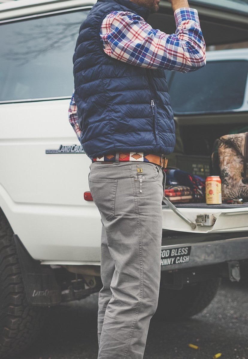 A person in a plaid shirt, blue vest, and gray pants stands by an open SUV trunk, wearing the Willie belt by Zilker Belts—an eye-catching hand-stitched Argentine leather accessory—with a drink can and bag inside.