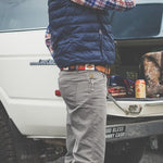 A person in a plaid shirt, blue vest, and gray pants stands by an open SUV trunk, wearing the Willie belt by Zilker Belts—an eye-catching hand-stitched Argentine leather accessory—with a drink can and bag inside.