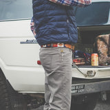 A person in a plaid shirt, blue vest, and gray pants stands by an open SUV trunk, wearing the Willie belt by Zilker Belts—an eye-catching hand-stitched Argentine leather accessory—with a drink can and bag inside.