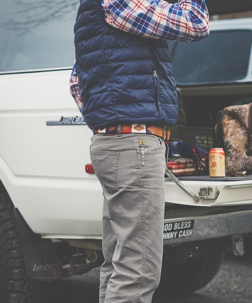 A person in a plaid shirt, blue vest, and gray pants stands by an open SUV trunk, wearing the Willie belt by Zilker Belts—an eye-catching hand-stitched Argentine leather accessory—with a drink can and bag inside.