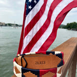 The Willie | Zilker Belt by Zilker Belts, hand-stitched Argentine leather with a southwest design and "ZILKER" embossed, rests on a wooden railing overlooking water and a U.S. flag.