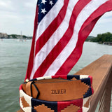 The Willie | Zilker Belt by Zilker Belts, hand-stitched Argentine leather with a southwest design and "ZILKER" embossed, rests on a wooden railing overlooking water and a U.S. flag.