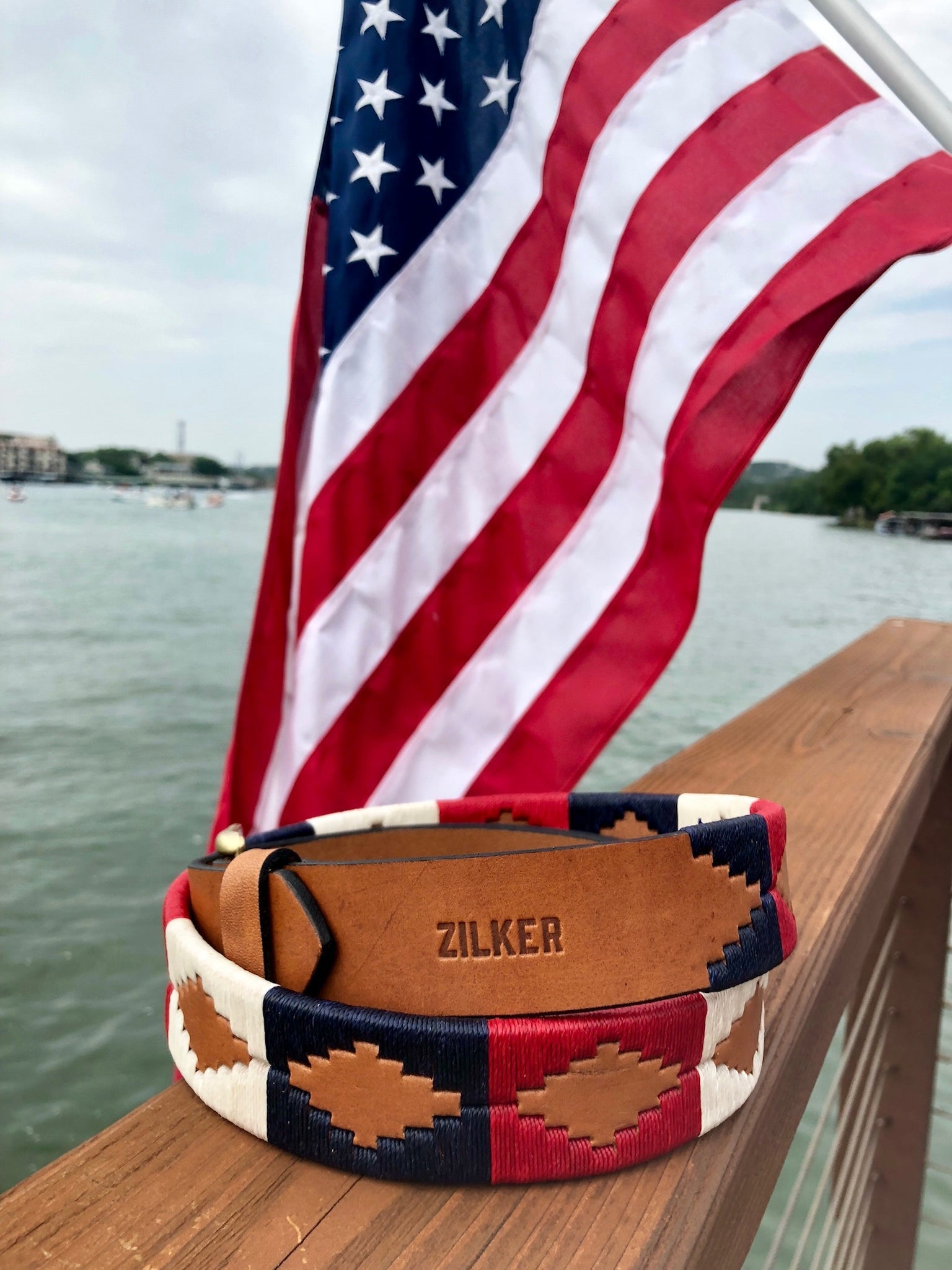 The Willie | Zilker Belt by Zilker Belts, hand-stitched Argentine leather with a southwest design and "ZILKER" embossed, rests on a wooden railing overlooking water and a U.S. flag.