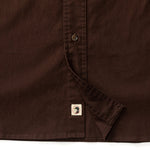 Close-up of the lower section of the Duck Head Woodland Shooting Shirt in brown stretch twill, featuring a button, stitched seam, and a small rectangular tag by the hem.