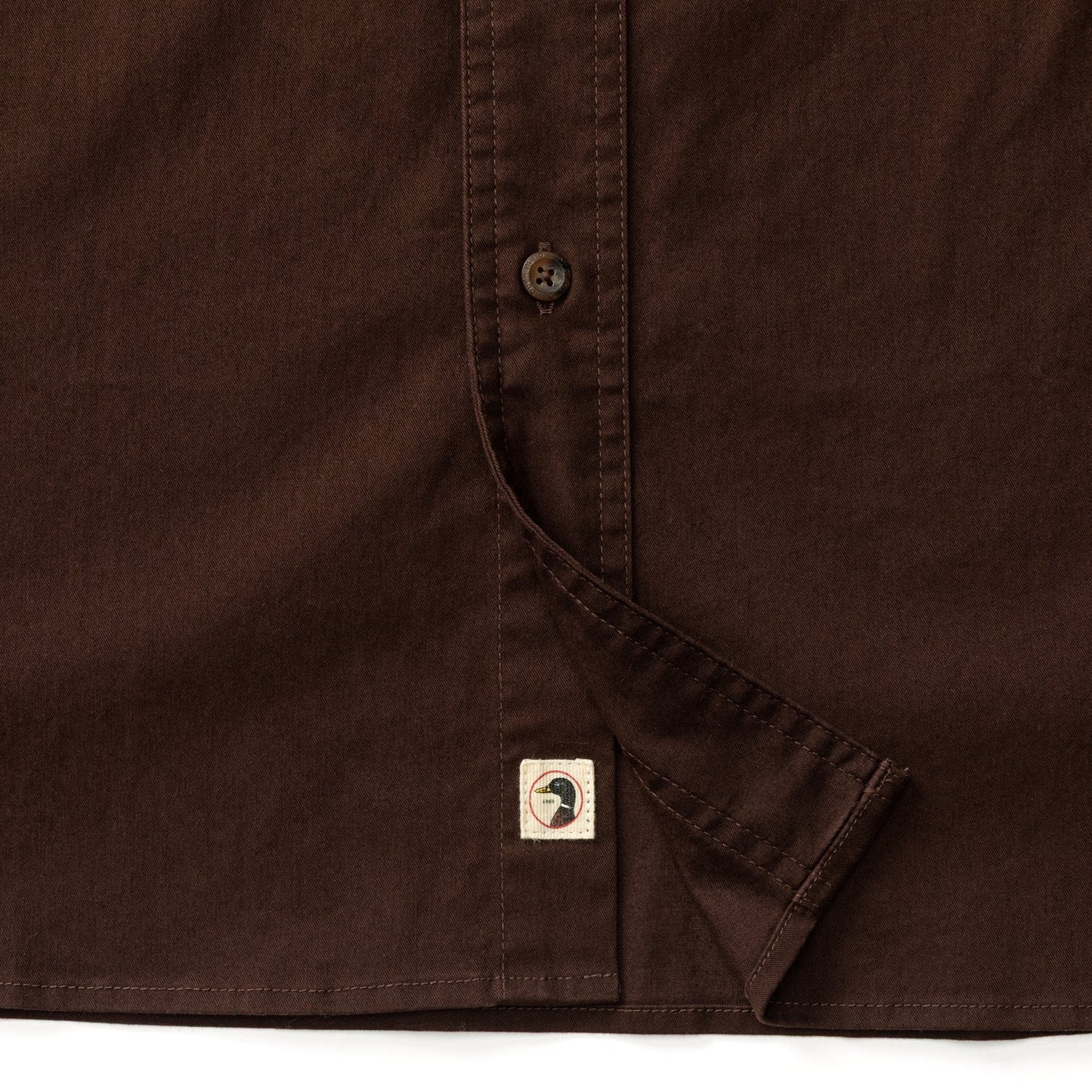 Close-up of the Duck Head Woodland Shooting Shirt, a brown stretch twill button-up featuring one visible button and a small fabric tag by the hem.