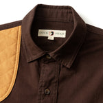 Close-up of the Duck Head Woodland Shooting Shirt in brown stretch twill, featuring a tan quilted shoulder patch for recoil protection and a signature "Duck Head" label inside the collar.