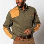 A man wearing the Duck Head Woodland Shooting Shirt—an olive green outdoor shirt with tan suede patches on the shoulders, elbows, and pocket—paired with brown pants and a belt.